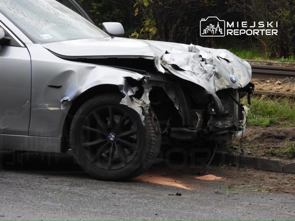 Uszkodzony samochód marki BMW z wgniecioną przednią częścią stoi przy drodze, obok torów kolejowych.