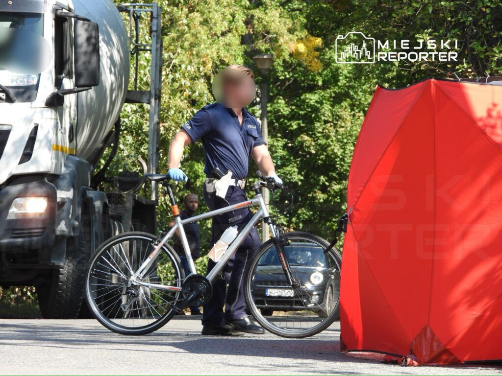 Policjant z rowerem zabezpiecza miejsce zdarzenia na drodze, obok stoi betonowy mixer.