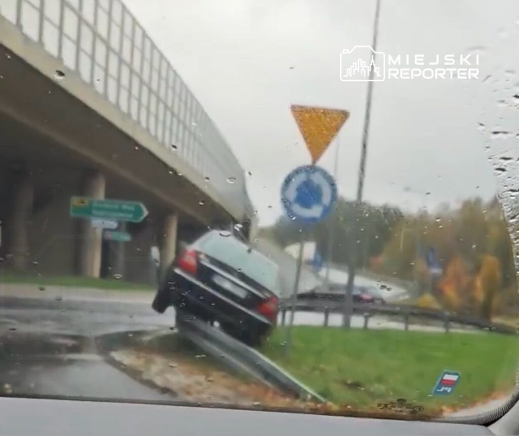 Samochód osobowy stoi na krawędzi drogi, z jednym kołem na trawie, obok znaku drogowym i pod wiaduktem.
