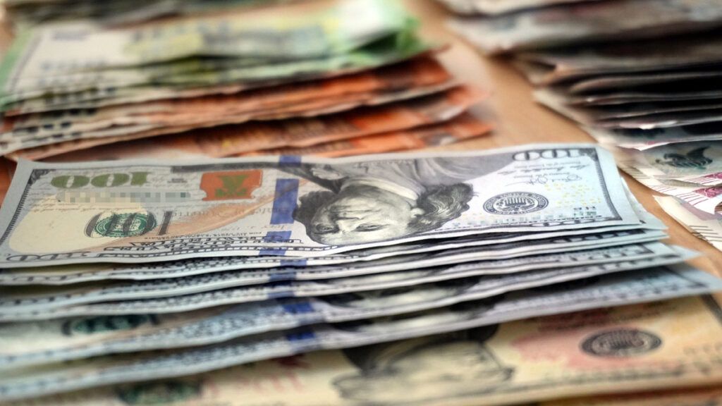 Stos banknotów różnych walut ułożony na drewnianym stole, w tym banknoty dolarowe i euro.