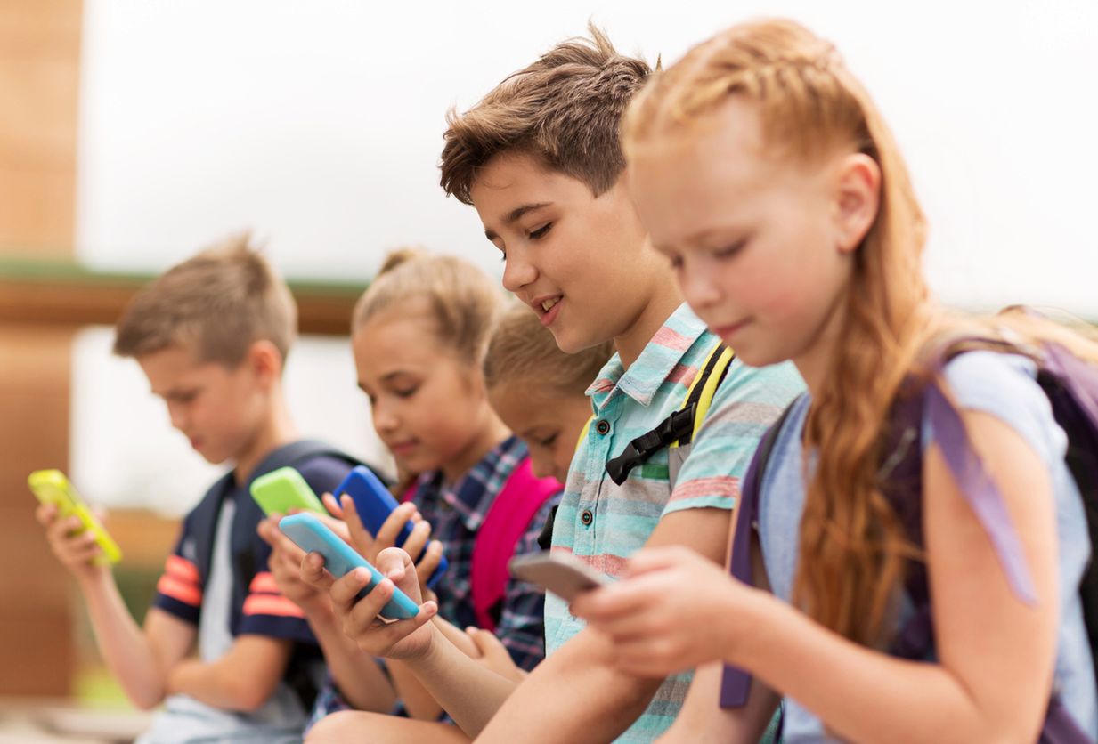 primary education, friendship, childhood, technology and people concept - group of happy elementary school students with smartphones and backpacks sitting on bench outdoors 