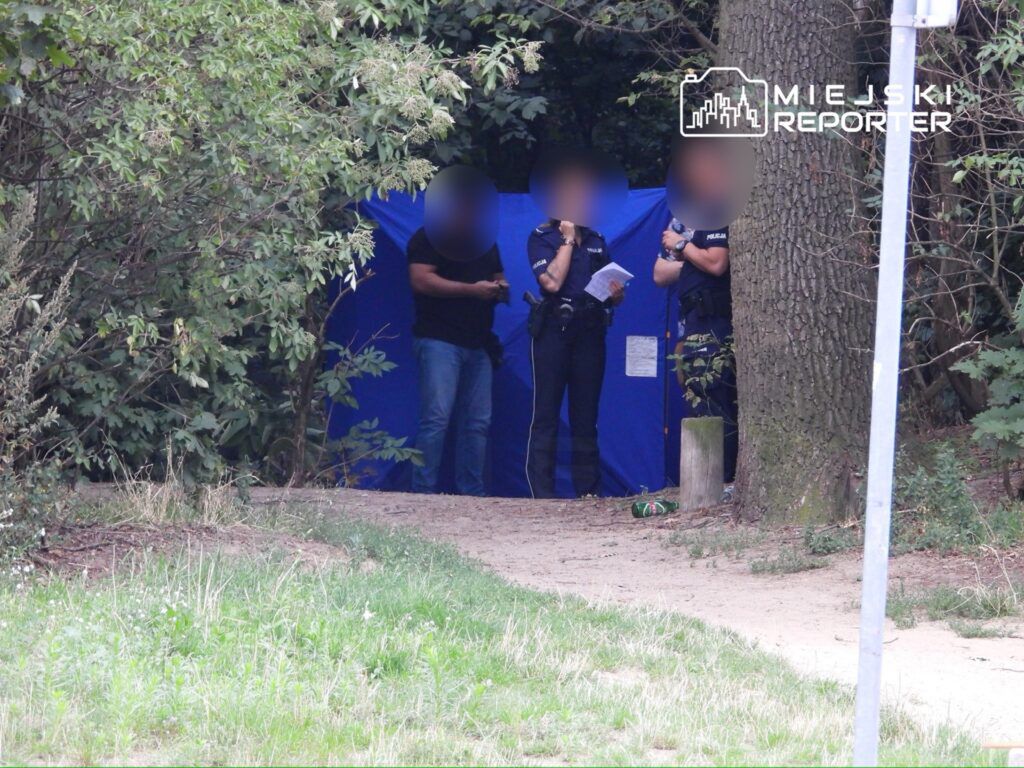 Grupa policjantów rozmawia w pobliżu niebieskiego namiotu w zalesionym terenie.