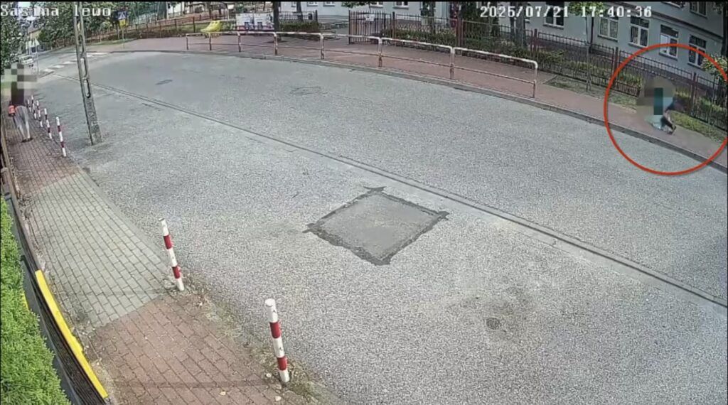 Mężczyzna w ciemnej odzieży spacerujący po chodniku przy ulicy, obok znaku drogowym i ogrodzeniu.