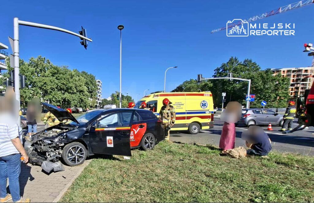 Czarny samochód osobowy z uszkodzoną maską stoi na chodniku, obok interweniują strażacy i karetka pogotowia.