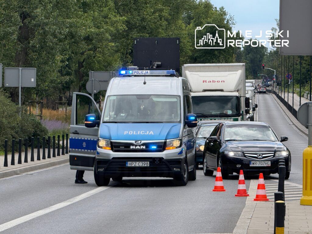 Policyjny radiowóz z otwartymi drzwiami kontroluje ruch drogowy, a w tle widoczne są samochody oraz sygnalizacja świetlna.