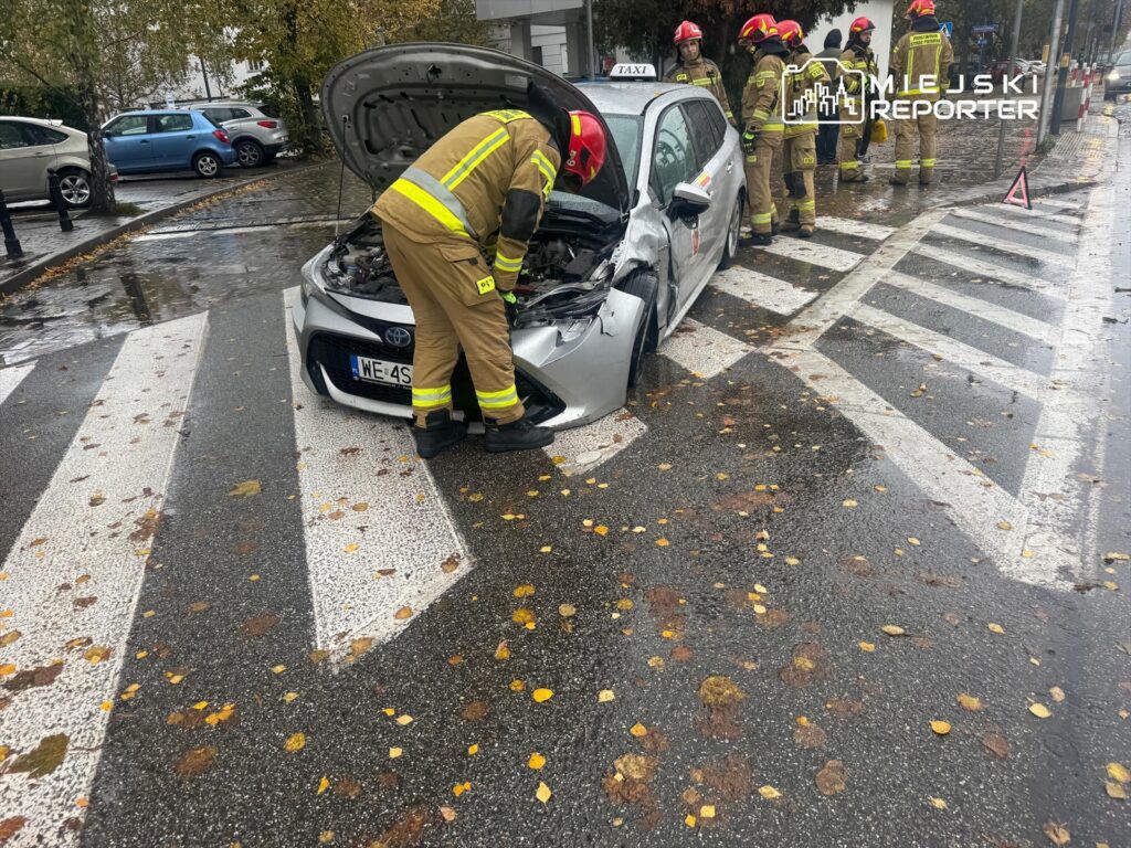 Strażacy sprawdzają uszkodzony samochód osobowy na pasach w deszczowej aurze, obok leżą liście.