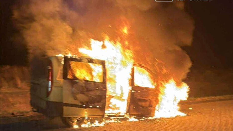 Zaparkowany pojazd płonący na ulicy, otoczony gęstym dymem i jasnym ogniem w nocy.