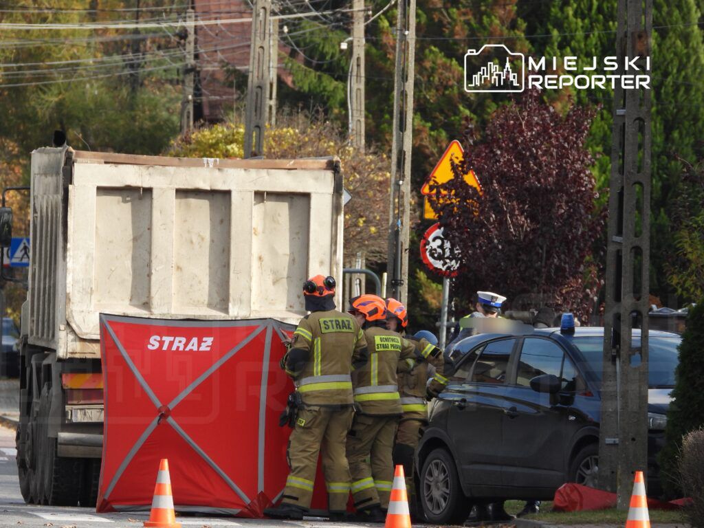 Strażacy w mundurach pracują przy czerwonym namiocie na ulicy, obok zaparkowanego radiowozu.