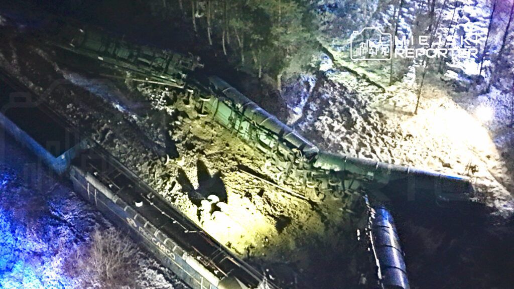 Złomowane wagony kolejowe leżą na boku w lesistym terenie, oświetlone nocnym światłem.