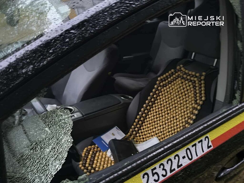 Zniszczona szyba samochodu osobowego, w środku widoczny fotel z drewnianym pokrowcem oraz dokumenty na siedzeniu.