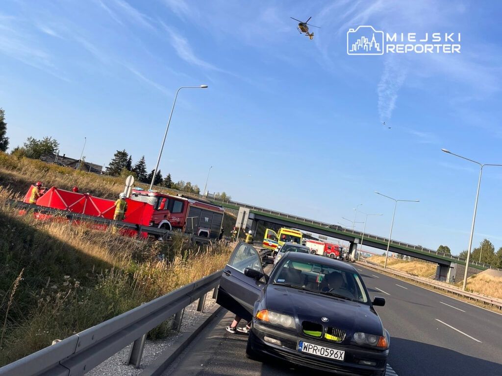 Na autostradzie, ratownicy w żółtych kamizelkach pracują obok czerwonego wozu strażackiego, a nad nimi krąży helikopter.