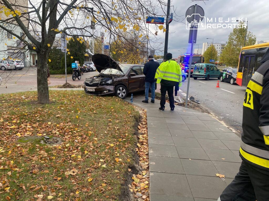 Samochód osobowy z otwartą maską stoi na trawie przy chodniku, obok grupy osób oraz pojazdów służb ratunkowych.