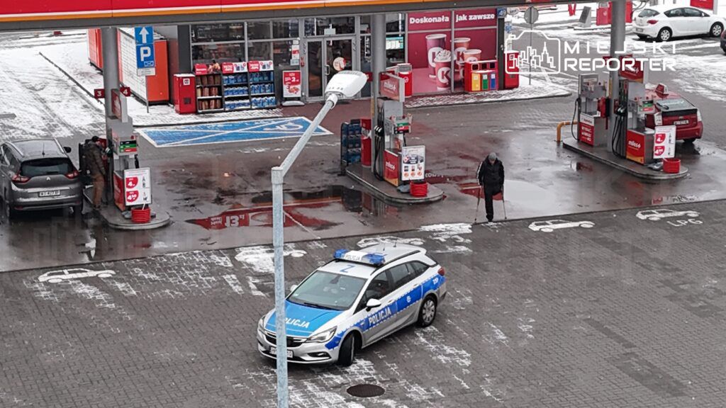 Mężczyzna tankujący samochód na stacji paliw, obok policyjny radiowóz, w tle zadaszona część stacji.