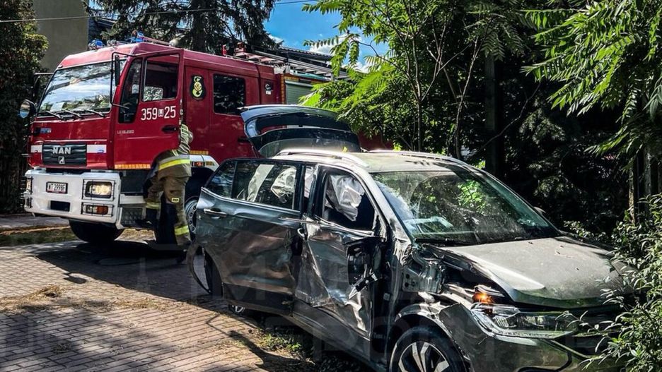 Strażak przy samochodzie osobowym z uszkodzeniami, obok widać wóz strażacki na osiedlowej ulicy otoczonej drzewami.