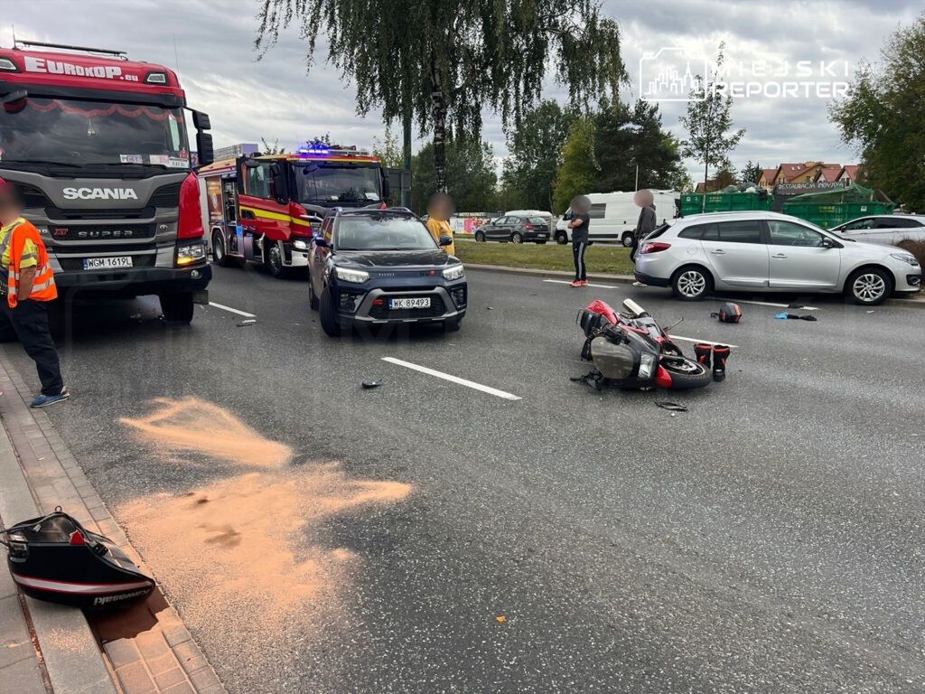 Na jezdni leży przewrócony motocykl, obok widać rozrzucone kaski i ślady piasku, a w tle stoją samochody i straż pożarna.