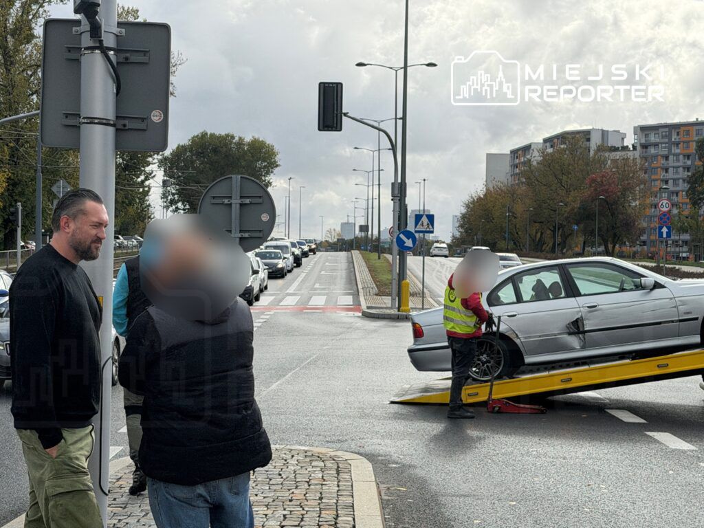 Mężczyzna w czarnym swetrze rozmawia z inną osobą obok drogi, podczas gdy holownik podnosi uszkodzony samochód.