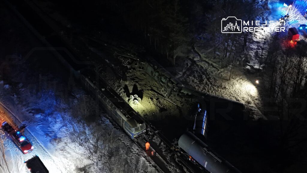 Zaniedbany pociąg towarowy leży na bocznicy, a w tle widać pojazdy ratunkowe oraz oświetlenie w lesie.