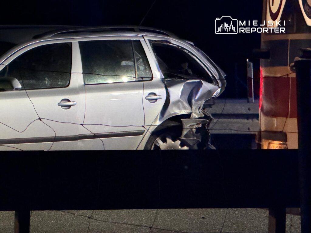 Uszkodzony samochód osobowy z wgniecioną tylną częścią stoi na poboczu drogi, obok niego widać pojazd służb ratunkowych.