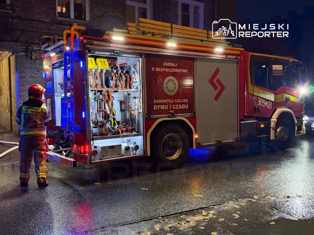 Strażak w mundurze przegląda wyposażenie w wozie strażackim na oświetlonej ulicy nocą.