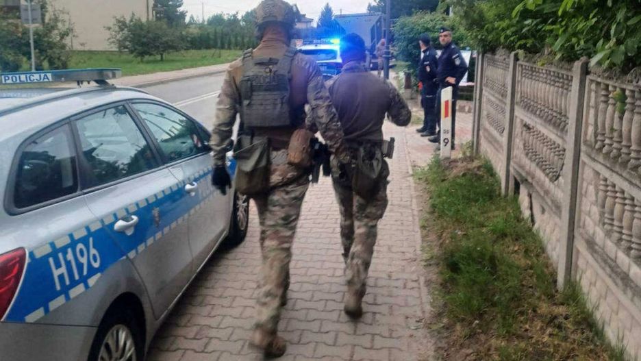 Dwóch mężczyzn w mundurach wojskowych idzie chodnikiem obok radiowozu policyjnego w miejskim otoczeniu.