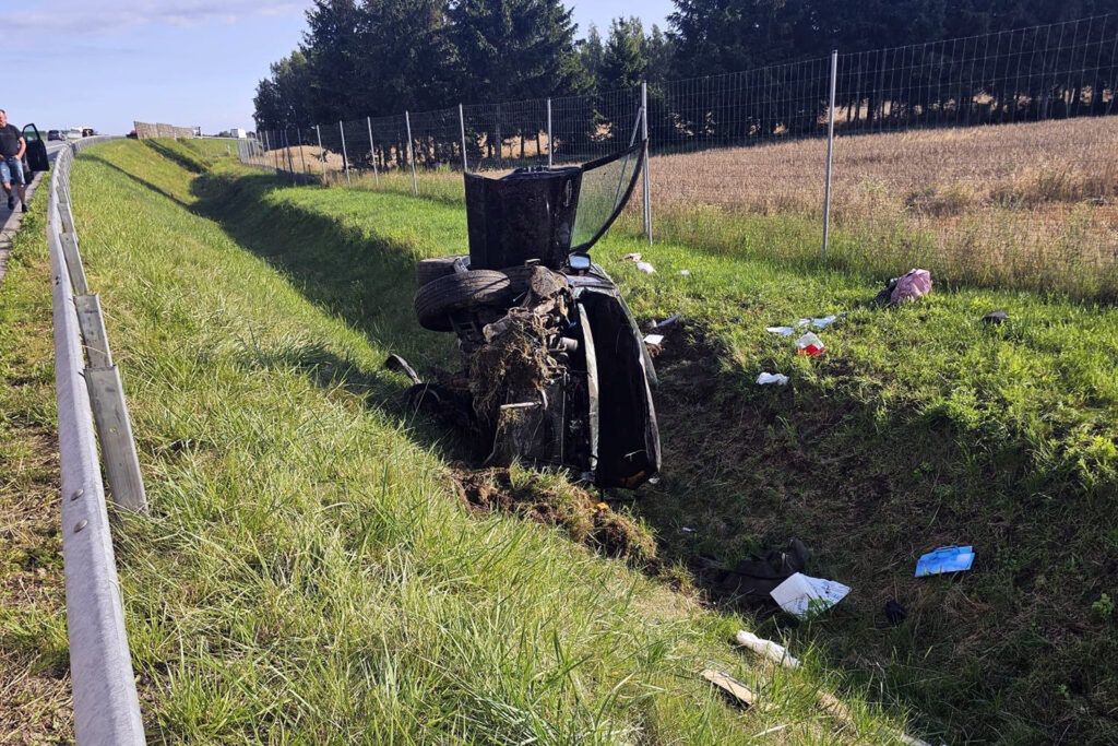 Zniszczony samochód leży na boku w rowie przydrożnym, obok trawy i odpadków. W tle widać drogę oraz drzewa.