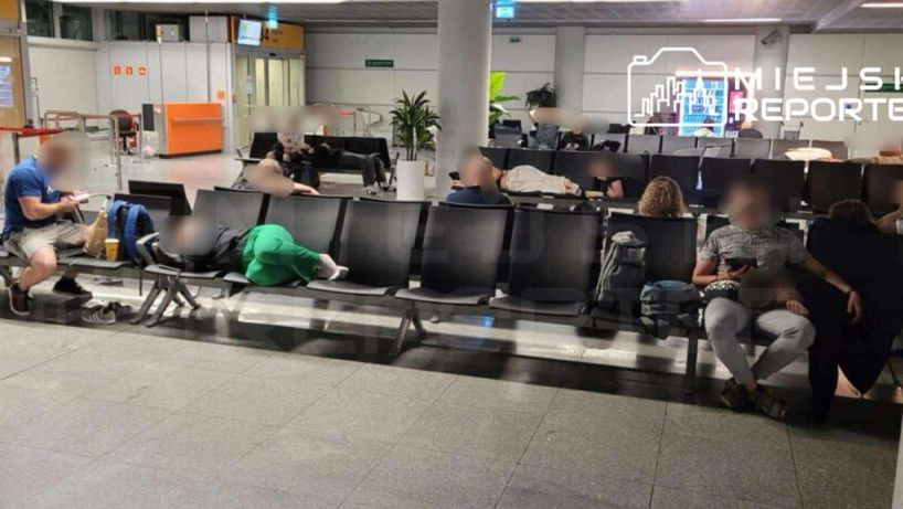 Grupa osób siedzi i leży na plastikowych ławkach w przestronnej poczekalni. W tle widać stanowiska odprawy.
