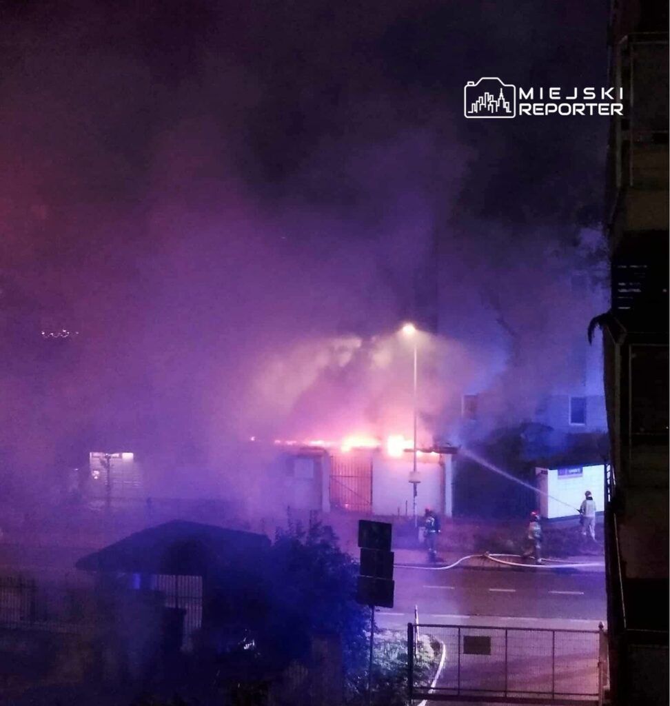 Ognisko pożaru otoczone gęstym dymem, w tle strażacy gaszący płomienie w nocy.