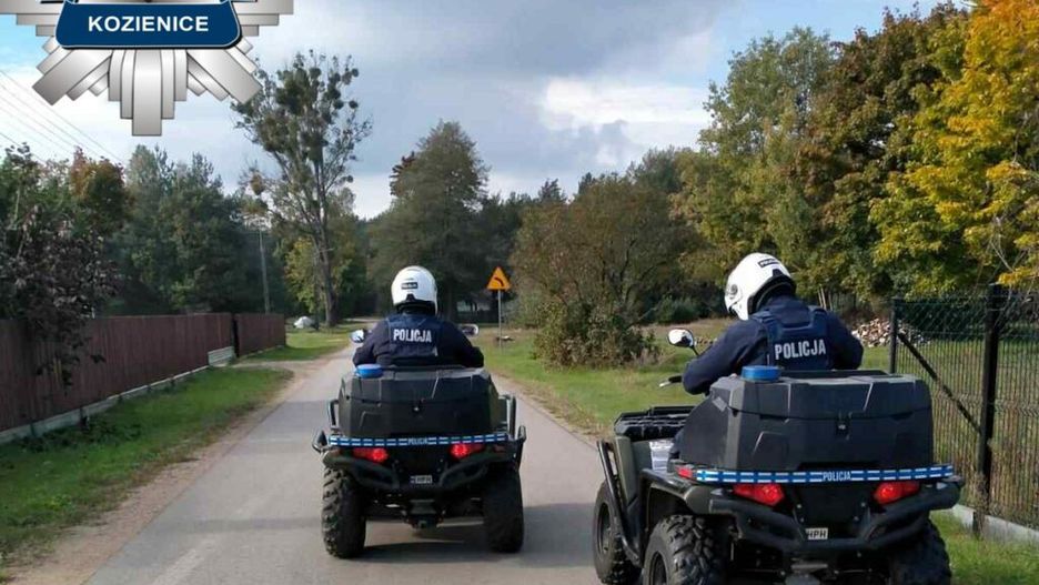 Dwaj policjanci na quadach patrolują wiejską drogę w otoczeniu drzew i ogrodzeń.