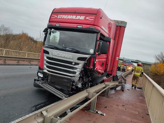 Czerwony samochód ciężarowy z uszkodzoną przednią częścią stoi na moście, obok pracują strażacy.