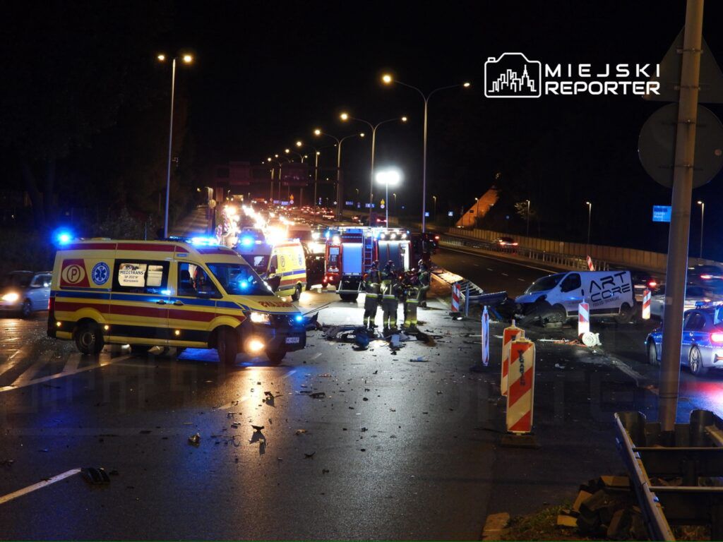 W nocy na drodze widać wypadek drogowy z uszkodzonymi pojazdami, wokół pracują strażacy i karetki.