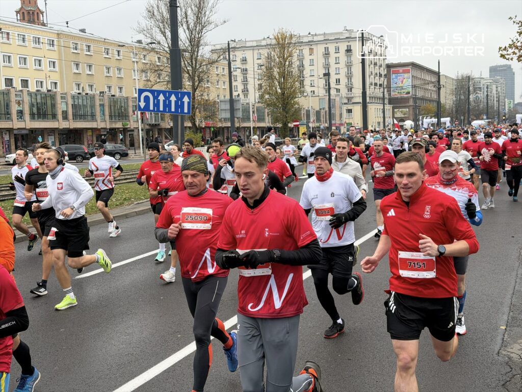 Biegacze w czerwonych koszulkach uczestniczą w wyścigu na ulicach miasta, mijając budynki i znaki drogowe.