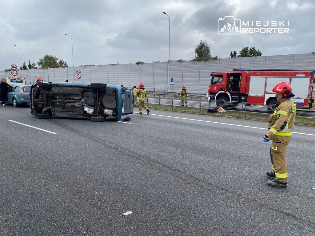 Na drodze leży przewrócony samochód, obok którego pracują strażacy w hełmach i mundurach. W tle widać inne pojazdy oraz woz strażacki.