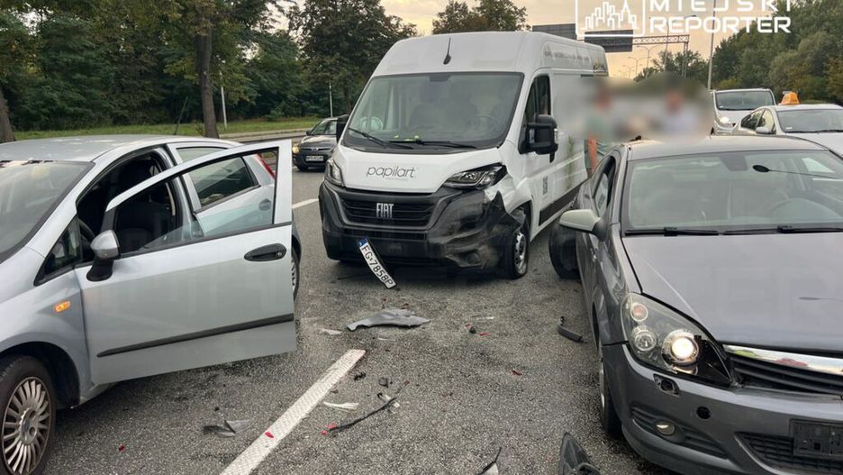Trzy uszkodzone pojazdy na drodze, w tym furgonetka z logo, z otwartymi drzwiami i odłamkami na jezdni.