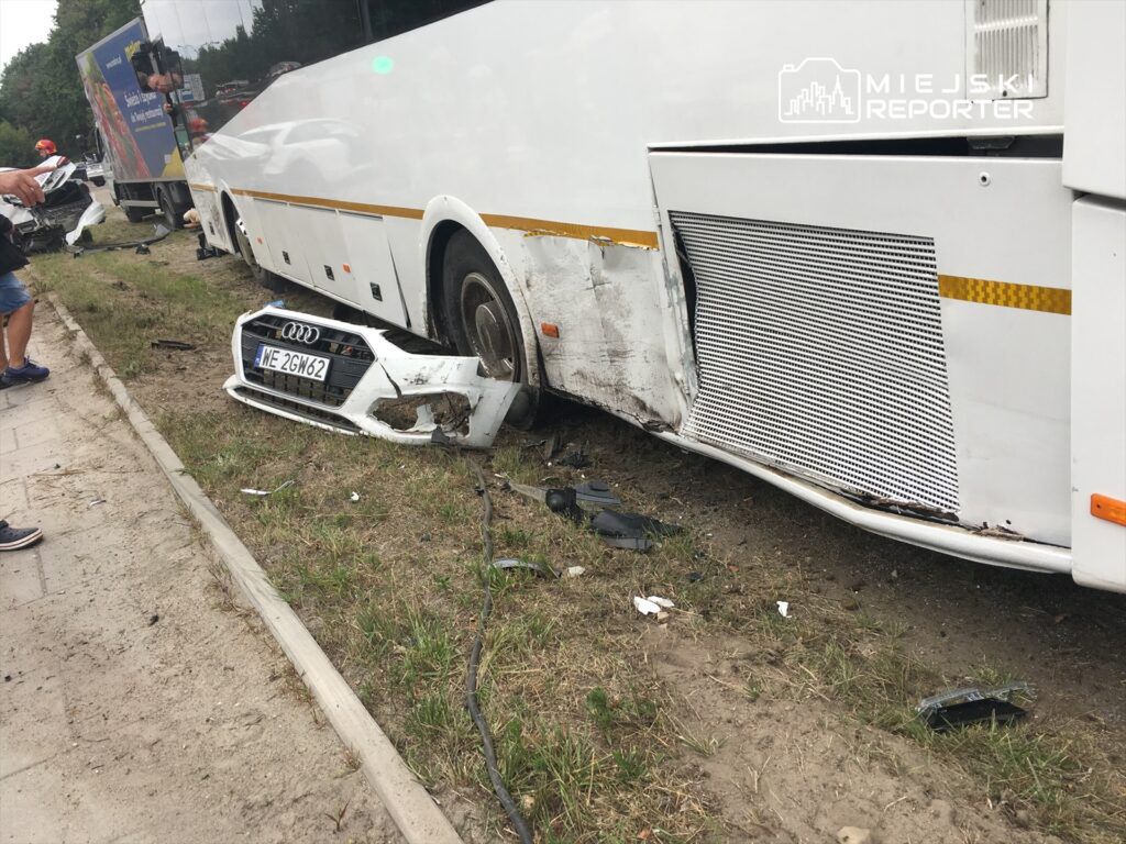 Zniszczony przód białego samochodu osobowego leży obok uszkodzonego autobusu na poboczu drogi.