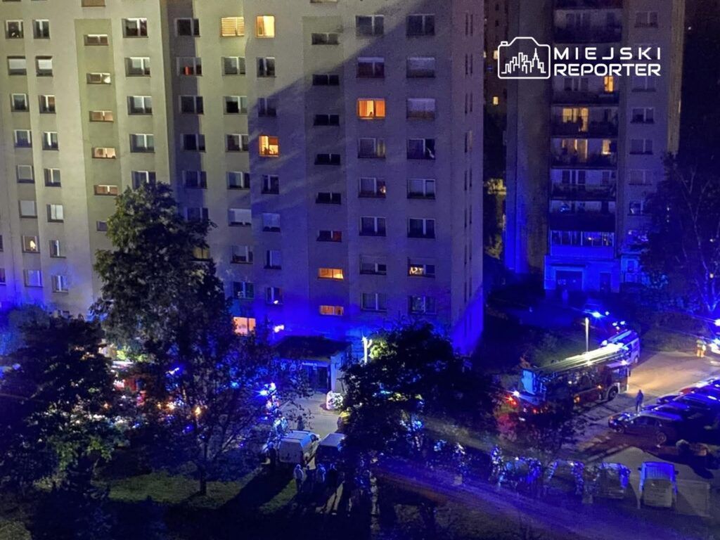 Grupa ludzi zbiera się w pobliżu budynku mieszkalnego, a w tle widać samochód strażacki z włączonymi sygnałami świetlnymi.