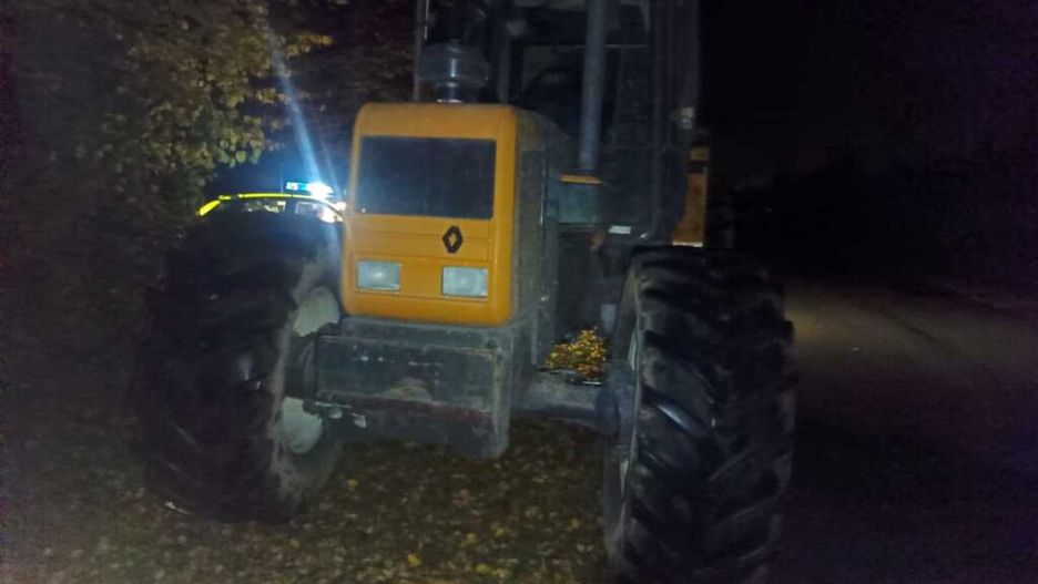 Traktor z pomarańczową kabiną stoi na drodze w nocy, w tle widoczna niebieska sygnalizacja.