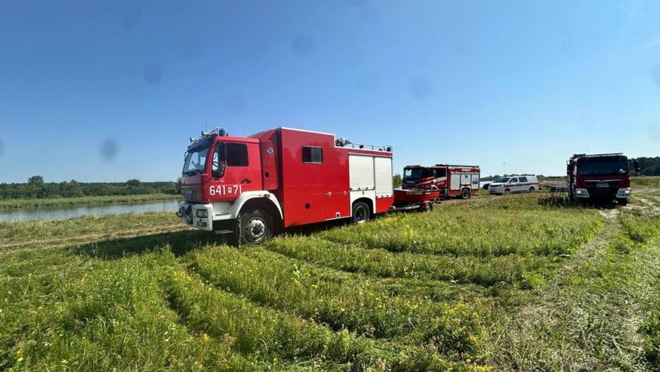 Czerwony wóz strażacki stoi na łące obok rzeki, w otoczeniu zielonej trawy i drzew.