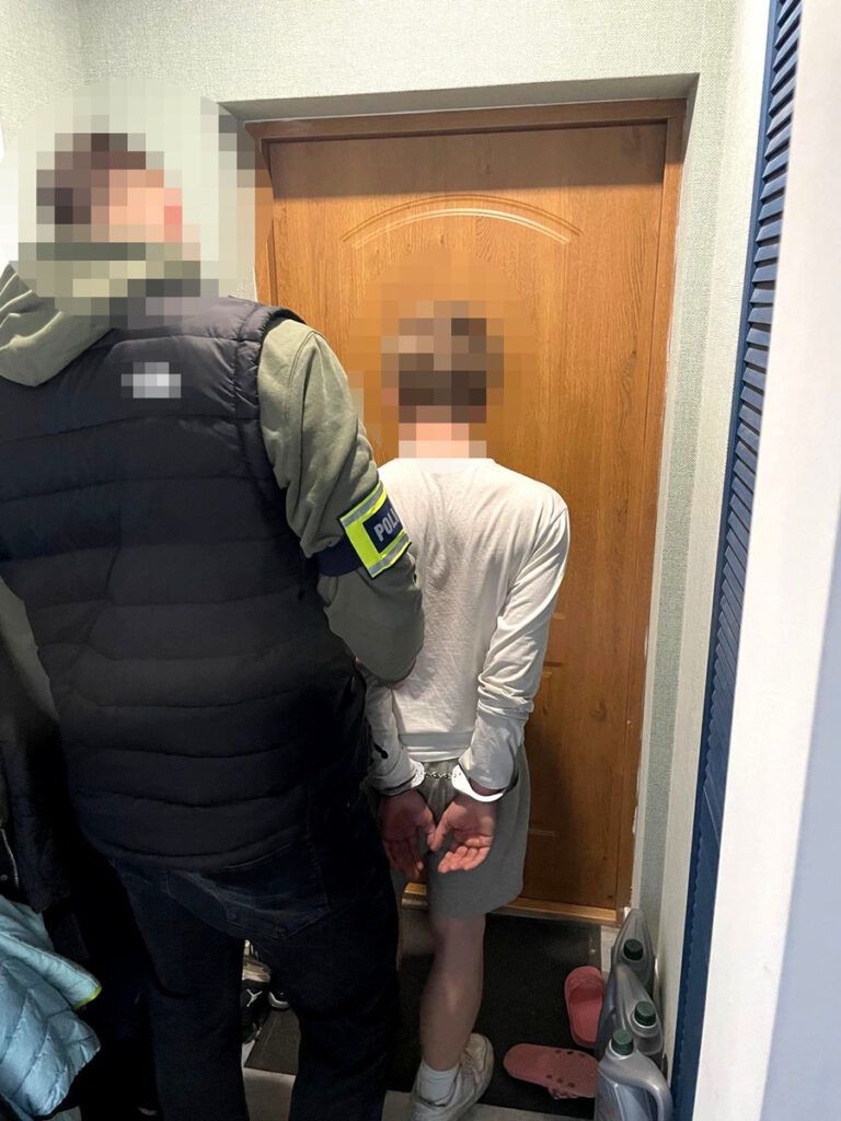 Mężczyzna w kajdankach stoi przy drzwiach, trzymany przez policjanta w kamizelce. W tle widoczne przedmioty domowe.