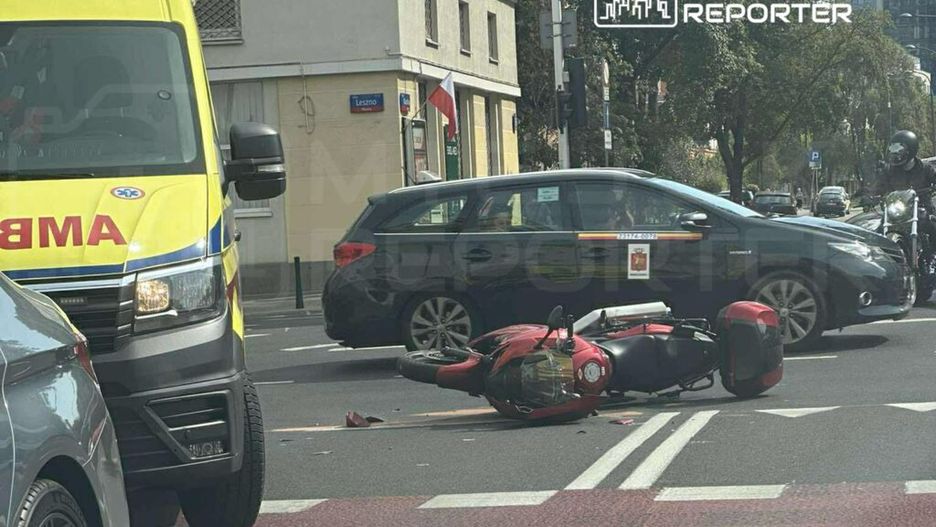 Ambulans zatrzymany przy przewróconym skuterze na ulicy, obok przejeżdżające samochody.