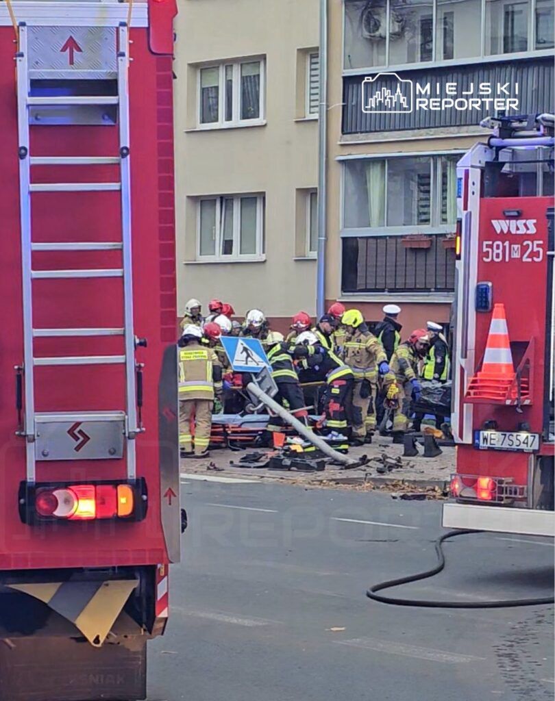 Grupa strażaków w hełmach i mundurach pracuje przy usuwaniu skutków zdarzenia na ulicy, obok wozu strażackiego.