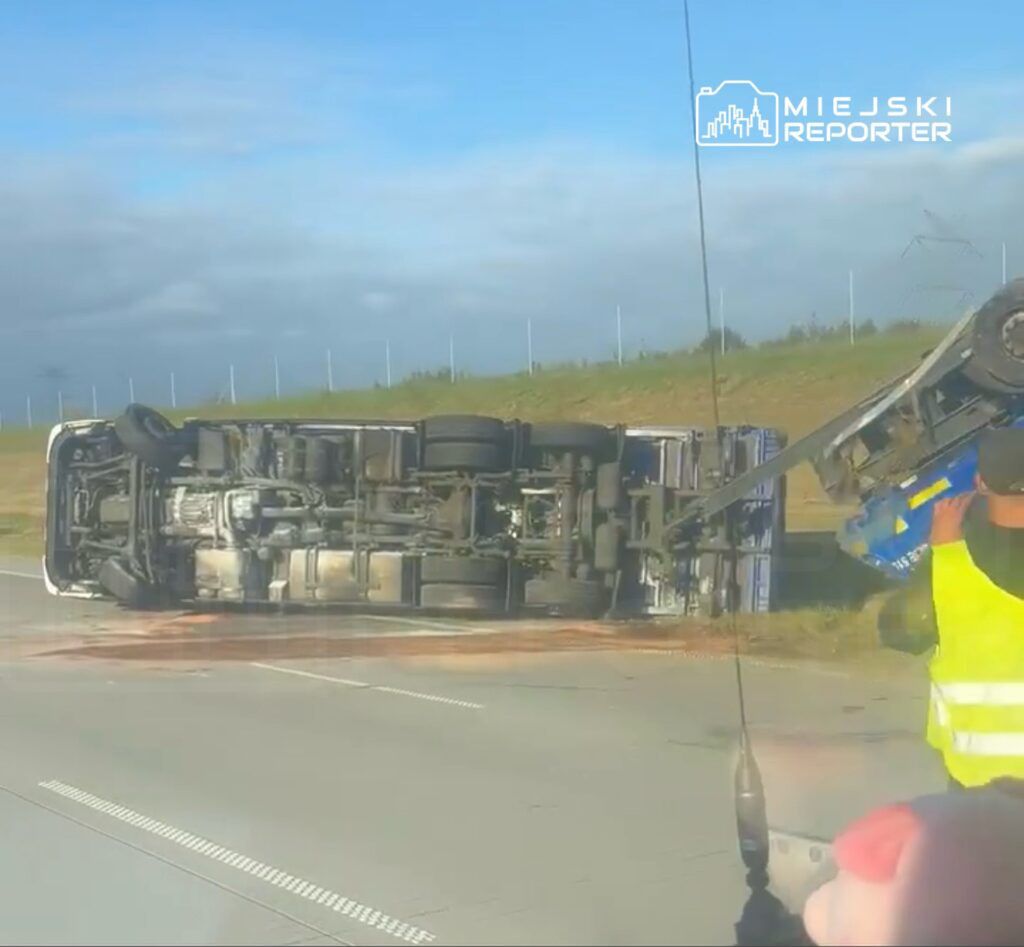 Ciężarówka leży na boku na drodze, a pracownik w kamizelce odblaskowej obserwuje akcję ratunkową.