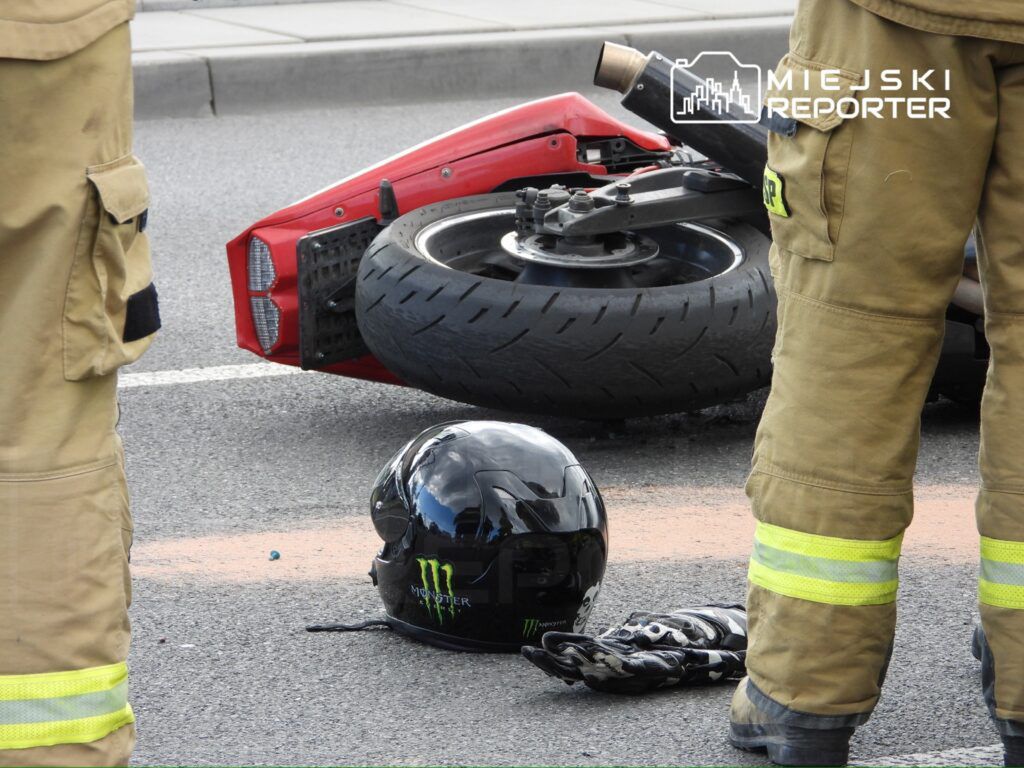 Motocykl leży na boku na drodze, obok niego czarny kask i rękawice, a w tle stoją strażacy w uniformach.