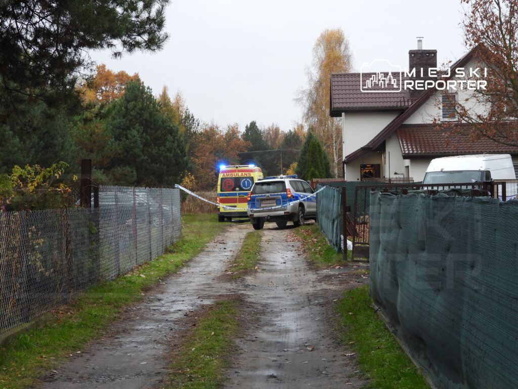 Policyjny radiowóz i karetka stoją na wąskiej drodze w otoczeniu domów i drzew, w tle widoczna jest siatka ogrodzeniowa.