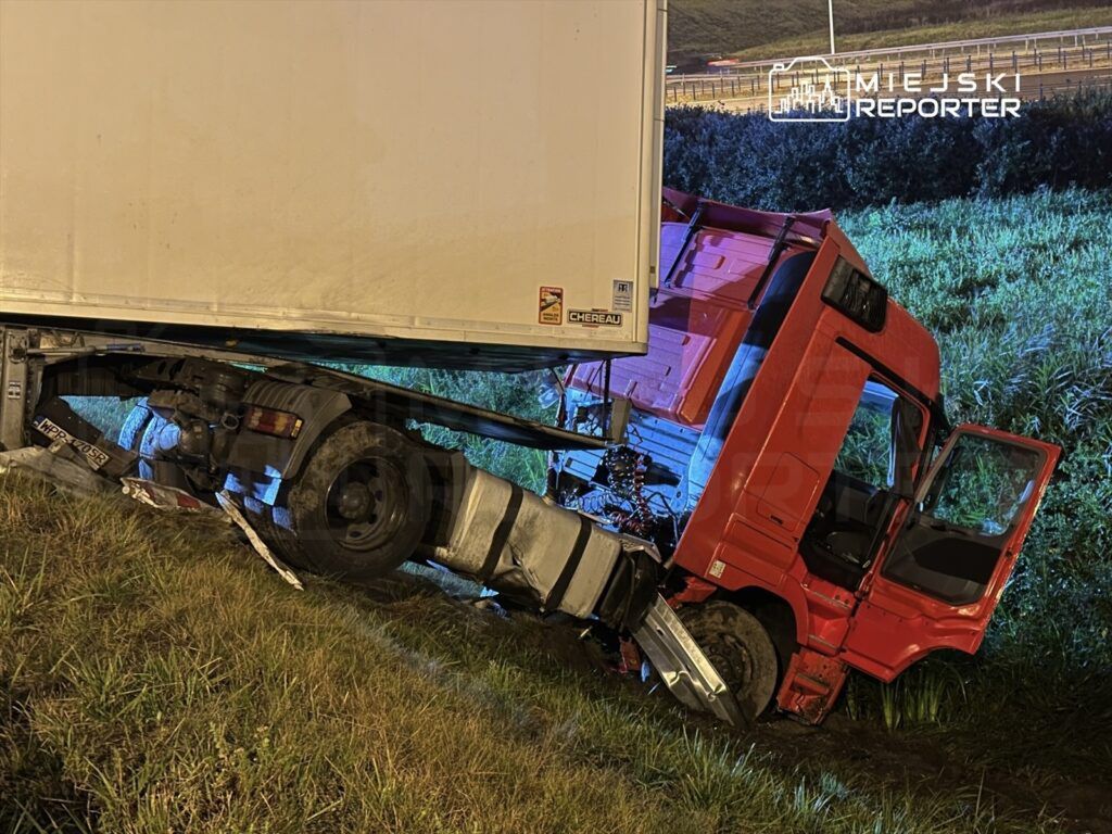 Czerwony ciężarówka z naczepą osunięta na pobocze drogi, część kabiny zanurzona w trawie.