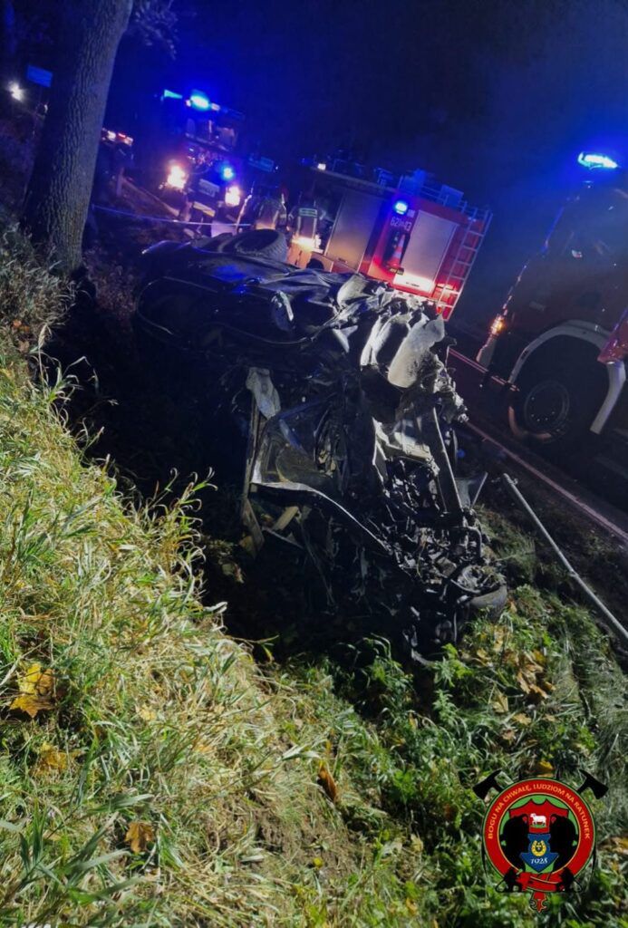 Wrak samochodu leżący na poboczu drogi w nocy, w tle widać pojazdy straży pożarnej z włączonymi sygnałami świetlnymi.