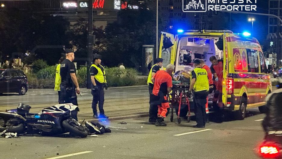 Na ulicy ratownicy medyczni transportują poszkodowanego na noszach do karetki, obok leży przewrócony motocykl.