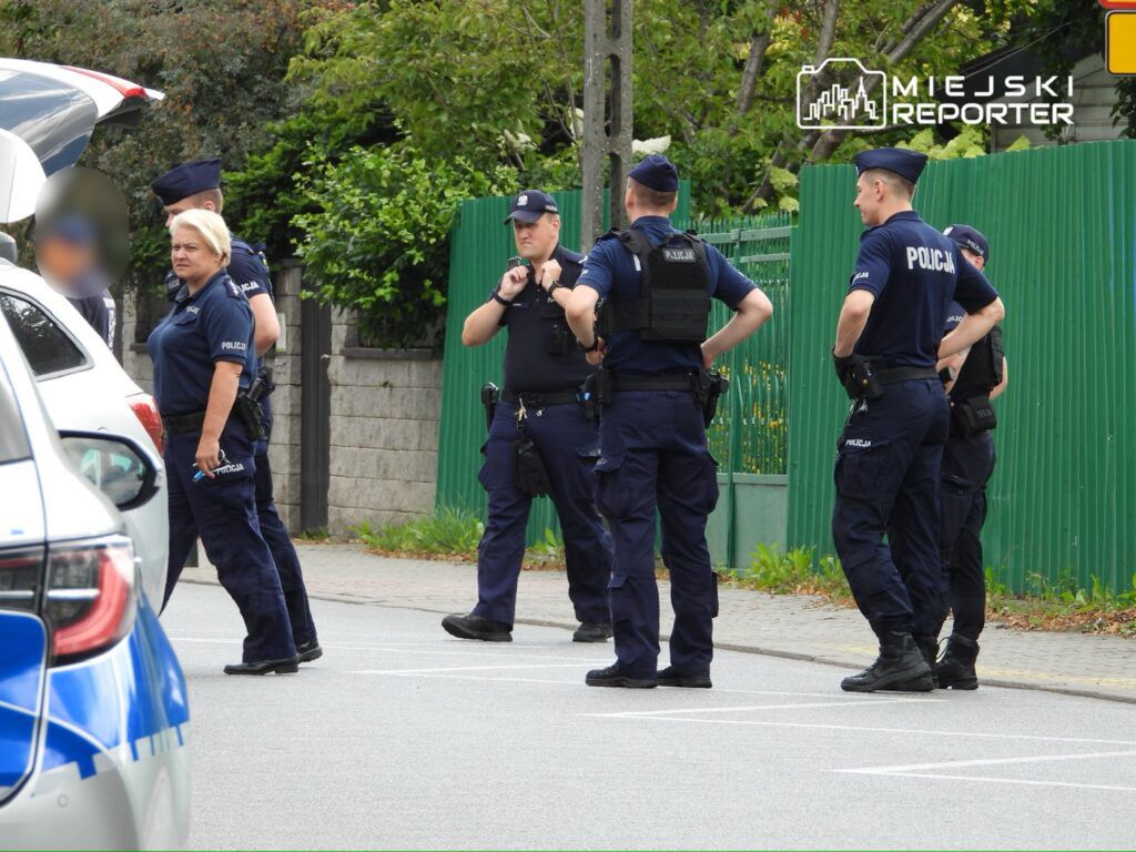 Grupa policjantów w mundurach stojąca na ulicy, prowadząca rozmowę w pobliżu zaparkowanego samochodu.