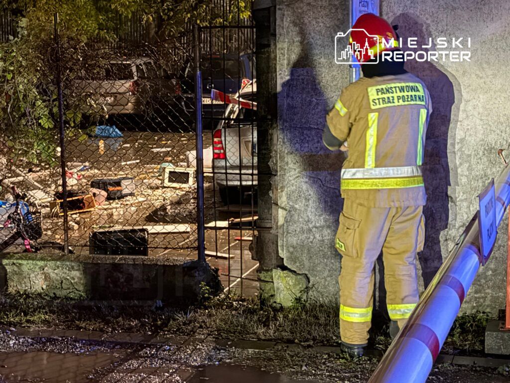 Strażak w mundurze z odblaskowymi elementami bada teren w zagraconym podwórku, w tle widać pojazdy i odpady.