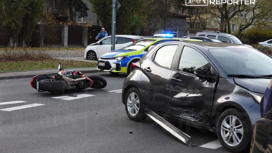 Motocykl leży na boku obok uszkodzonego samochodu osobowego na ulicy, a w tle widać radiowóz policyjny.