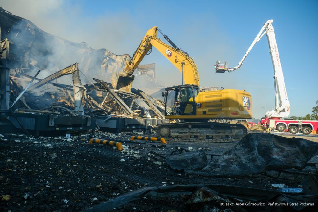 Żółty koparka rozbiera zniszczony budynek w otoczeniu dymu i zgliszczy, obok widoczna drabina strażacka.